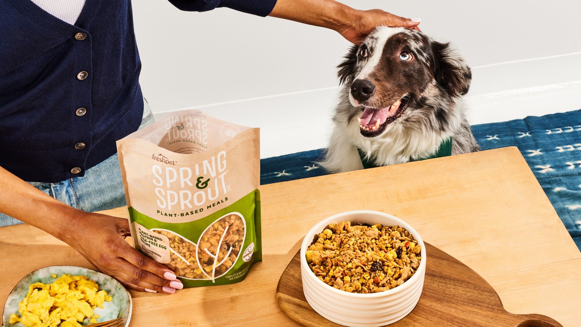 A black and white dog being pet with a bag and bowl of Freshpet Spring & Sprout in the foreground. 