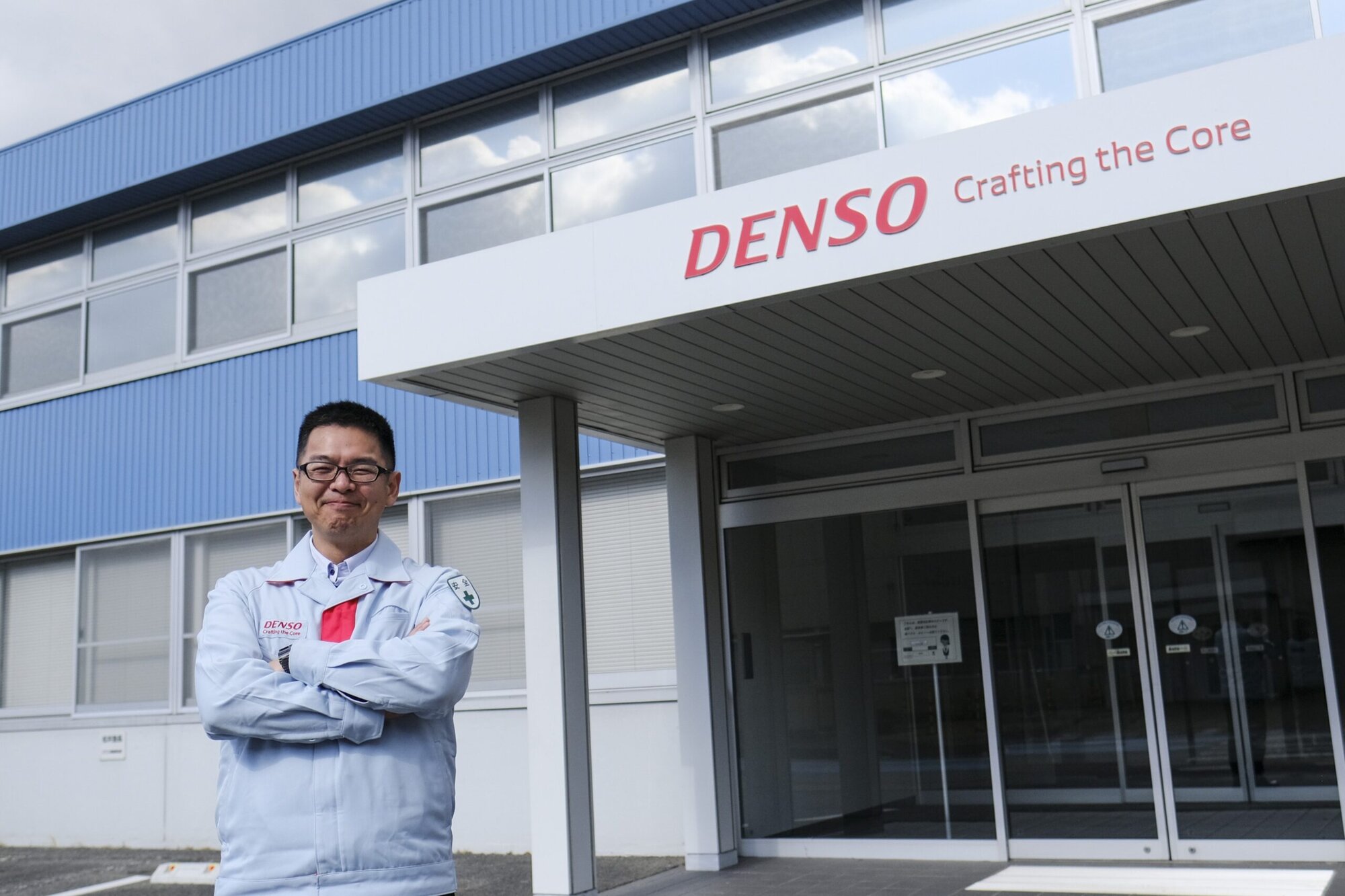 Takahiro Oba in a DENSO work jacket standing with arms crossed outside a DENSO building with the sign 'DENSO Crafting the Core'