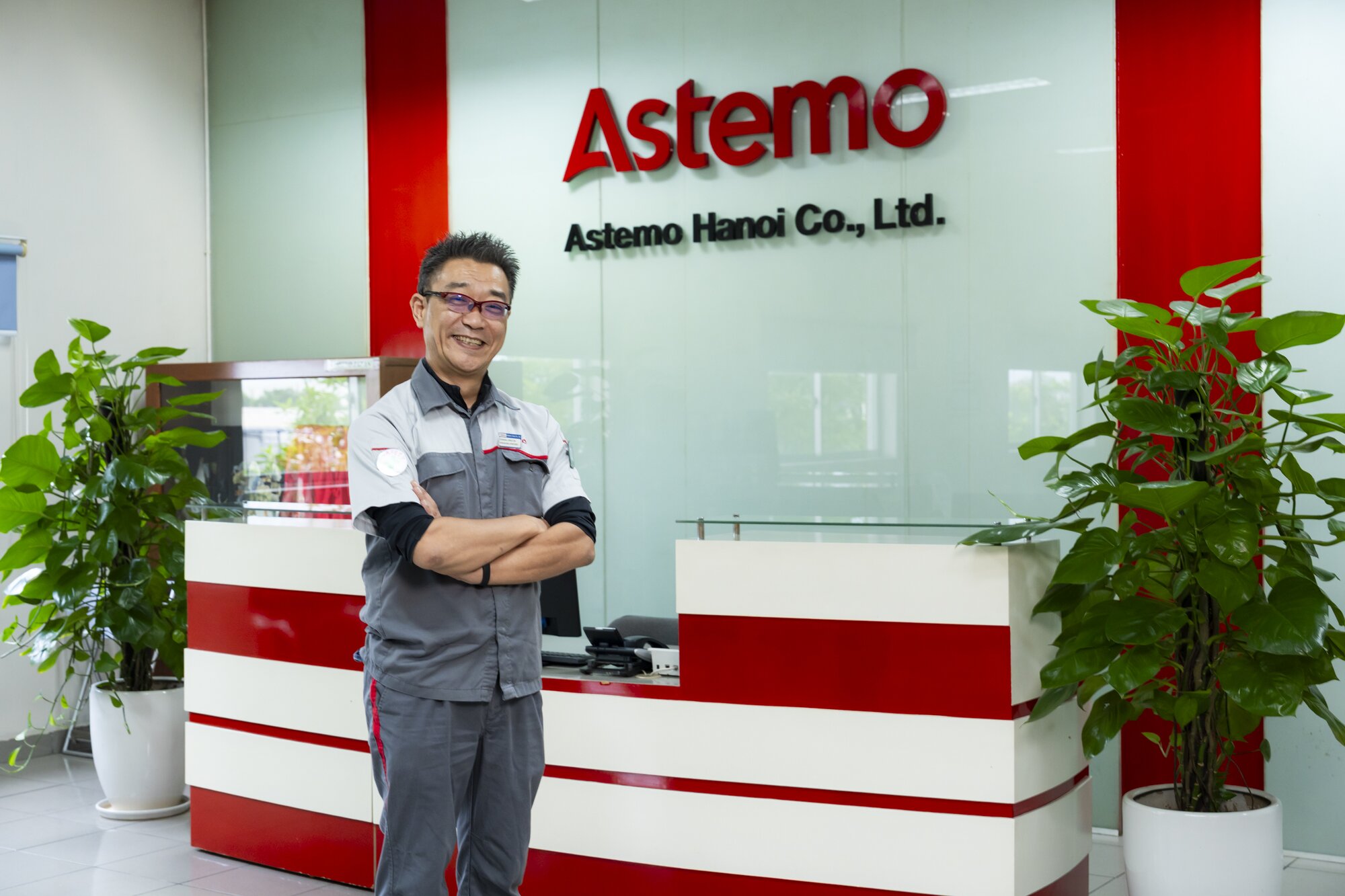 Smiling man in a gray work uniform standing with arms crossed in front of the Astemo Hanoi Co., Ltd. reception desk, flanked by potted plants.
