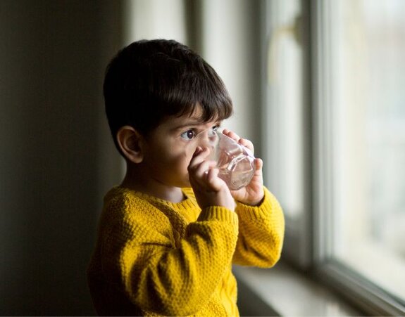 A young child drinks filtered, clean water from a glass while looking out of a window.