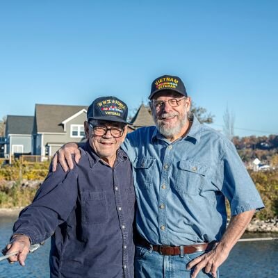 Two Vietnam veterans stand with their arms around one another's shoulders on a dock.