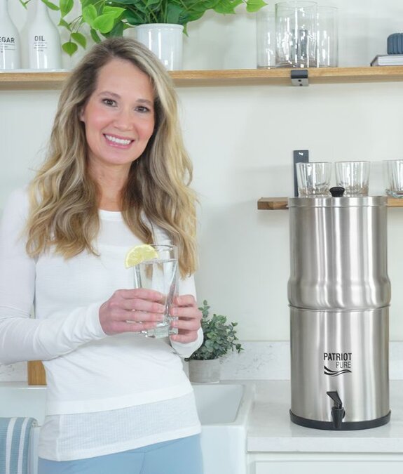 A woman in a white shirt in her kitchen holding a delicious glass of water filtered using the 4Patriots Ultimate Water Filtration System.