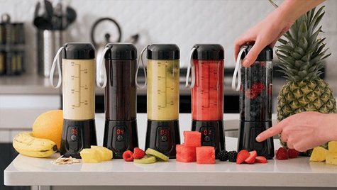 Five Patriot Power blenders on a counter next to a variety of fruit with someone turning on one of the blenders