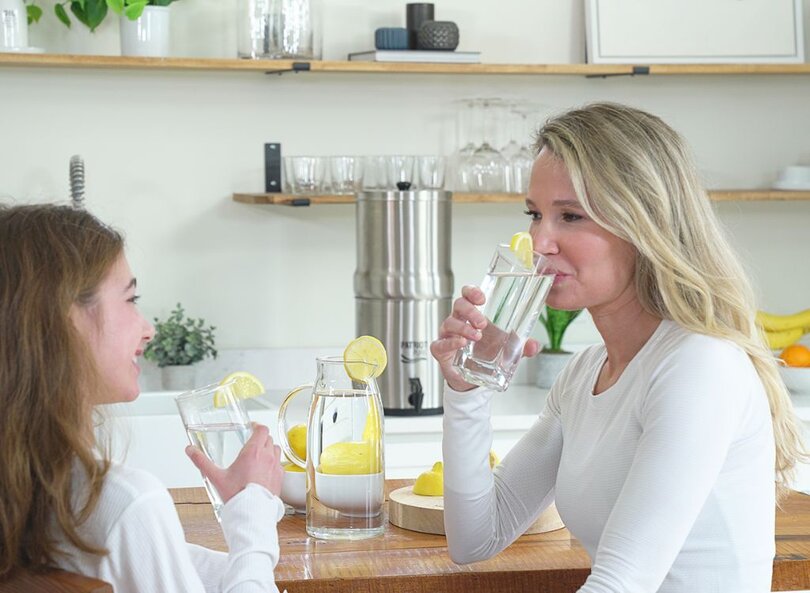 A mother and daughter drink clean filtered water in their kitchen with the Ultimate Water Filtration System on the counter.