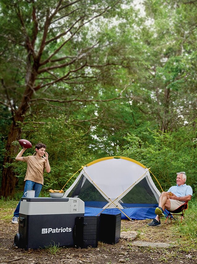 A grandpa, father, and son throwing a football in the woods next to their tent and their Freedom Fridge Cooler as they camp. 