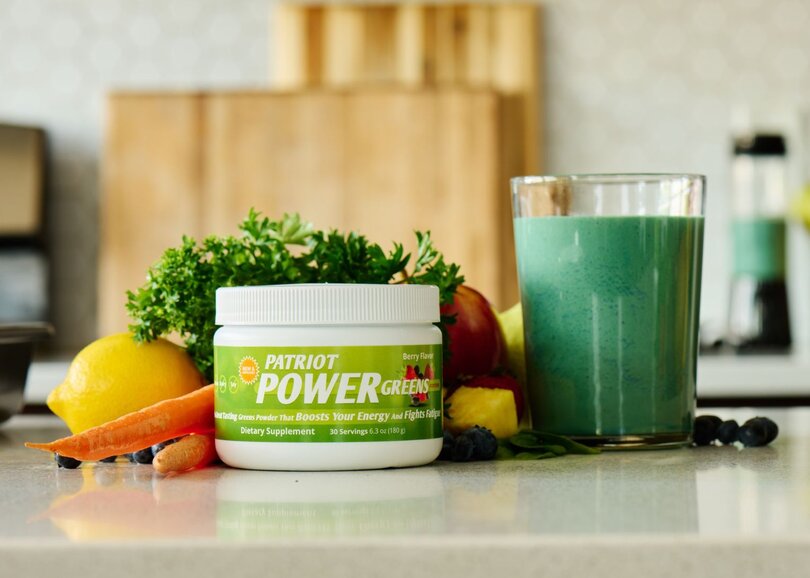 A canister of Patriot Power Greens on a kitchen counter next to a glass full of prepared Greens drink surrounded by fresh fruits and veggies.