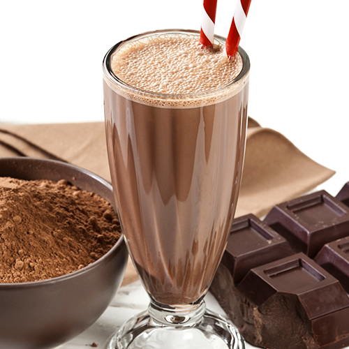 Chocolate survival shake in a glass with 2 straws next to a bowl of cocoa and a bar of chocolate.