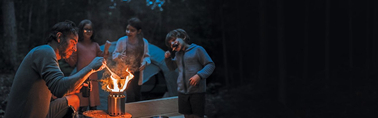 A family of 4 outside at night camping in the woods while they cook dinner on the StarFire Campstove