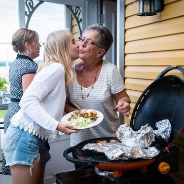 A teenage girl gives her grandmother a kiss on the cheek while the grandmother grills outside.
