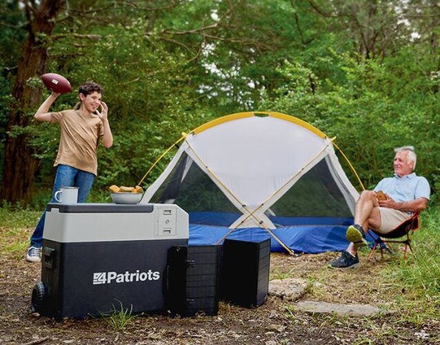 A child throws a football at a campsite with his grandfather and a 4Patriots Solar Go-Fridge in the front keeping their camp food cool.