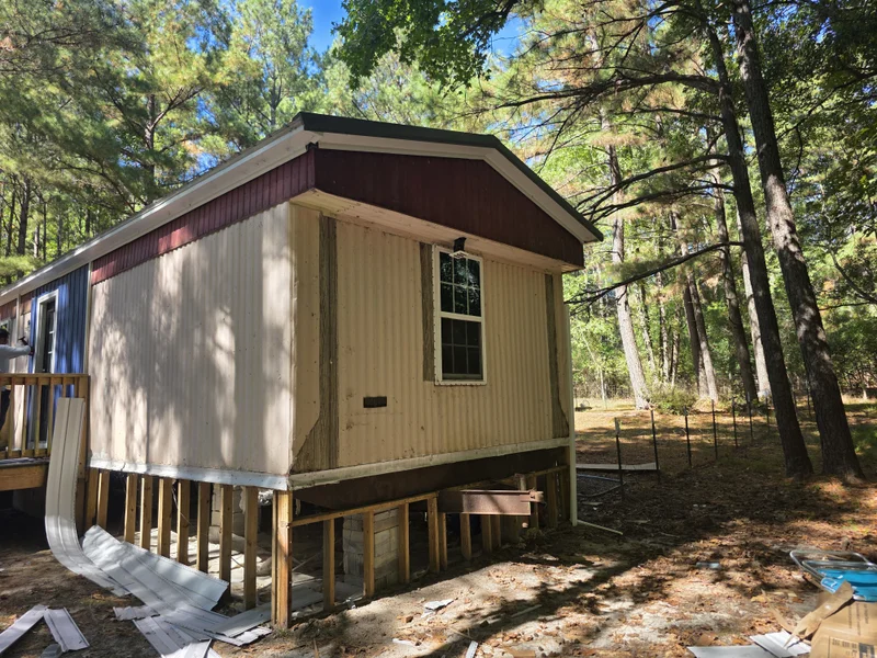 Manufactured home with red and tan metal siding before re-siding, surrounded by pine trees