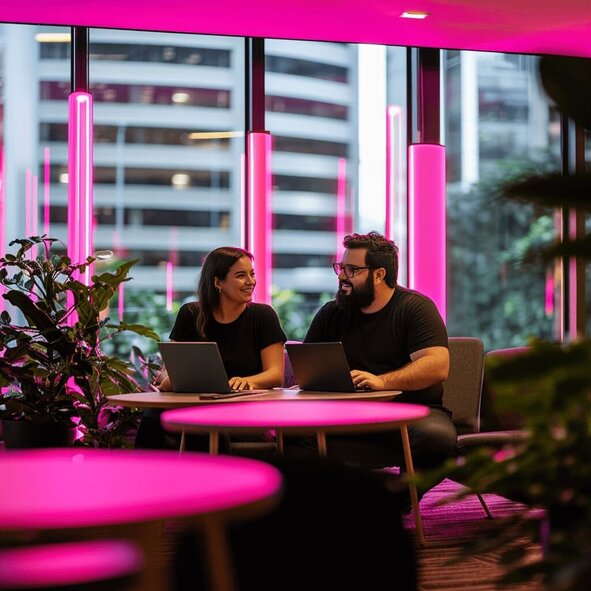 Homem e mulher sentados juntos. Ambos mexem em um notebook. Ao fundo, vê-se janelas de vidro com vista para prédios. Ilustrando o trabalho remoto de alguém que conta com um escritório virtual para endereço fiscal da empresa.