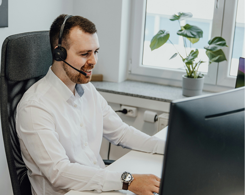 A man wearing a headset is seated at a desk, working intently on a computer in a modern office setting.