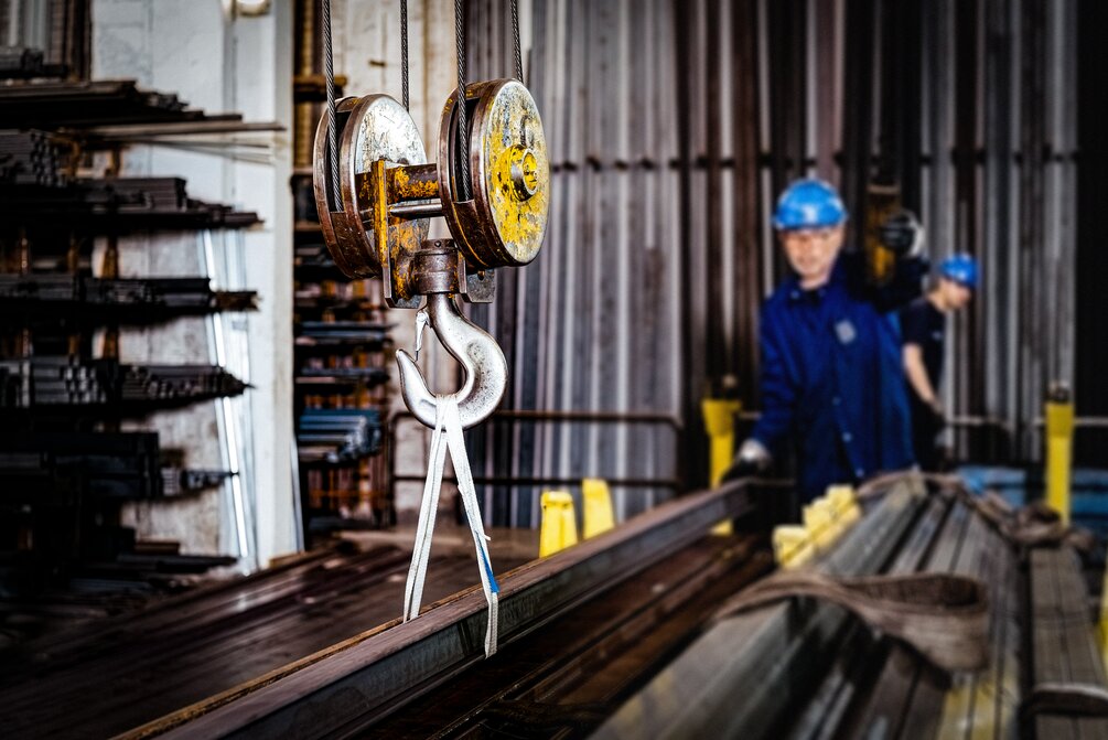 Industrial warehouse scene with a large metal hook suspended from a pulley system in the foreground, and two workers in blue uniforms and helmets handling metal beams in the background.