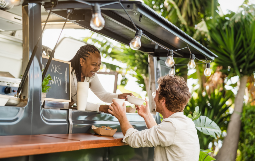 A food truck vendor hands a bowl and a cup to a customer at an outdoor counter, with a small chalkboard sign reading "TAKE AWAY," string lights above, and green plants in the background.