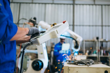 Person in a blue uniform holding a control device in an industrial workshop, with robotic arms and sparks from welding or cutting in the background.