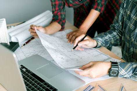 Two individuals collaborating over architectural blueprints at a desk, with a laptop, rolled-up plans, and drawing tools present.