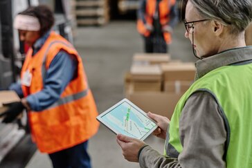 Warehouse scene with two workers in high-visibility vests; one holds a tablet showing a map or route, while the other handles boxes in the background