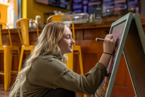 Customer handing an item to a barista wearing an apron behind a coffee shop counter, with coffee machines and equipment in the background.