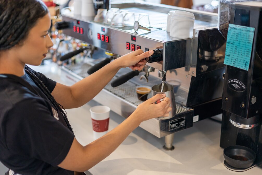 Person in a black apron standing behind a café counter with coffee dispensers and digital menu boards in the background, interacting with a customer.