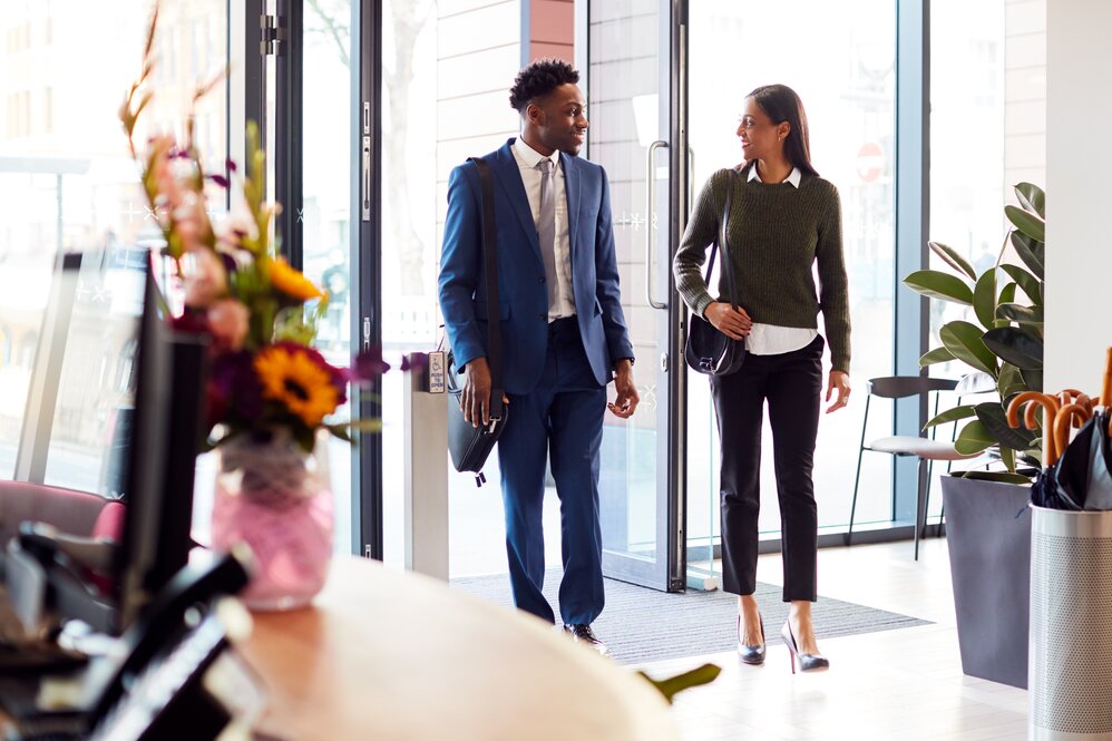 Two people enter a modern office lobby, one in a blue suit and the other in a green sweater and black pants, both carrying black bags.