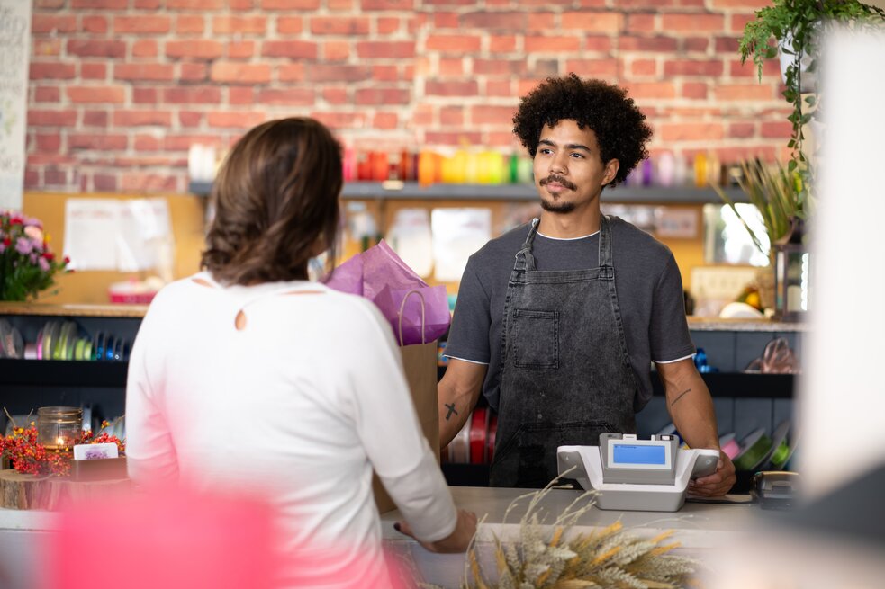 Florist wearing an apron standing behind a counter next to a point-of-sale terminal in a flower shop, with flowers, plants, and ribbons in the background.