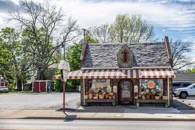 A waiter serves food to two people seated at a table in a restaurant with brick walls, large windows, and hanging light fixtures. Other diners are visible in the background.