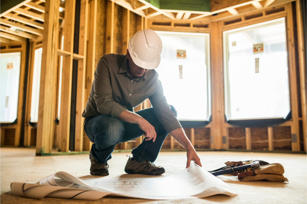 Person wearing a white hard hat and gray shirt kneeling on a construction site floor, examining and pointing to architectural blueprints, with wooden framing and large windows in the background.