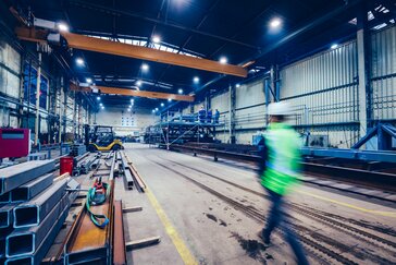 Interior of an industrial warehouse with metal beams, cranes, and heavy machinery. A worker in a high-visibility vest and hard hat walks through the space.