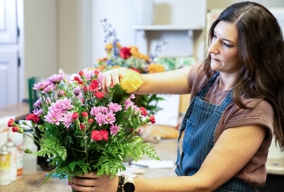 Florist wearing an apron standing behind a counter in a flower shop, with flowers, plants, and ribbons in the background.