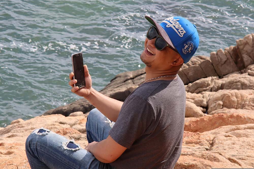 A happy man sitting on rocks beside the sea using his smartphone to apply for a line of credit.