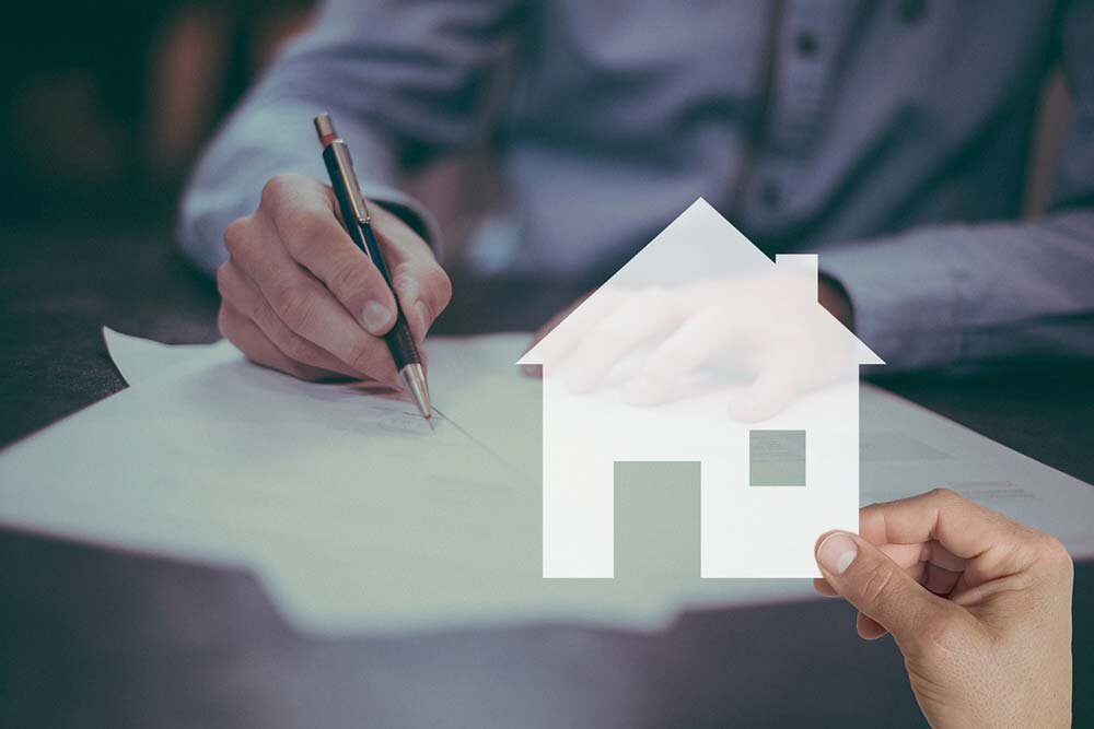 A man seated signing loan papers and another hand holding out a paper cut out of a house.