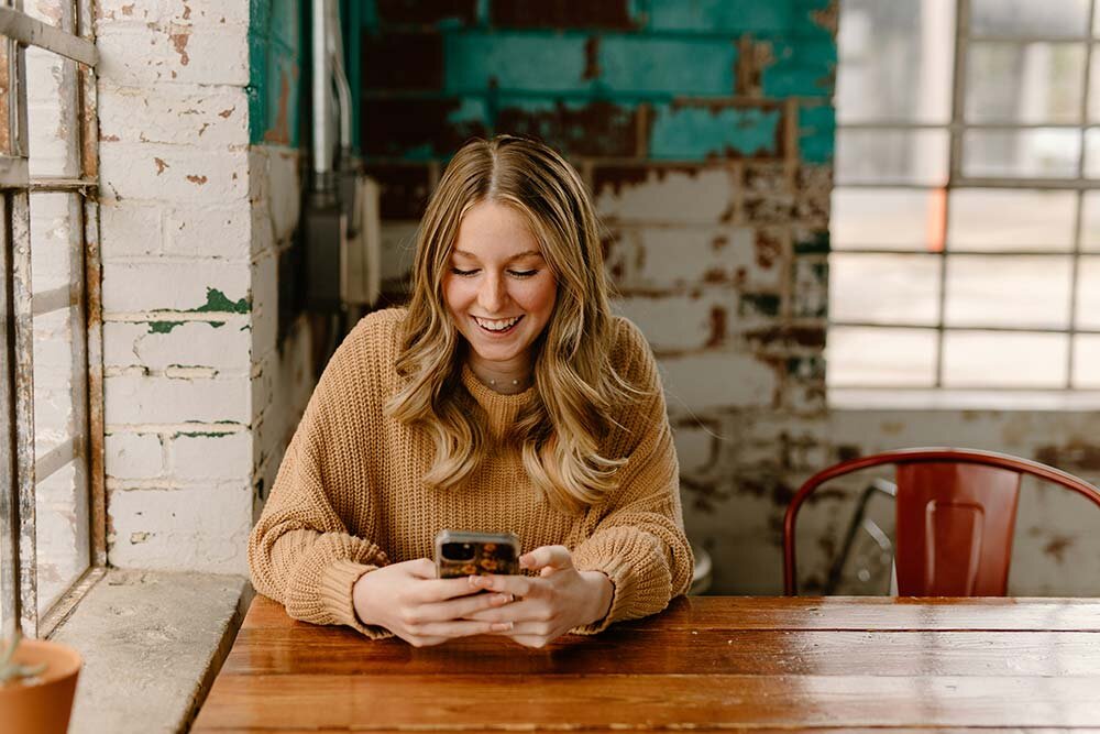 A happy woman setting at a table next to a window looking down a cell phone