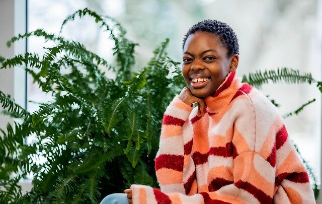 Smiling black woman sitting next to a large decorative fern