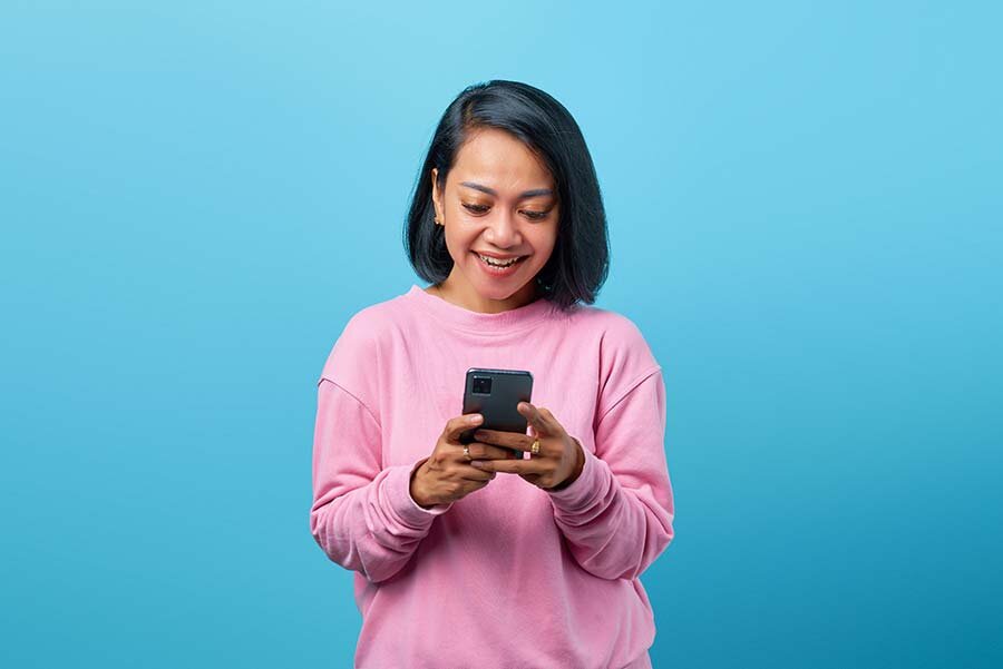 A happy woman in a pink shirt using a cellphone  to check her DTI ratio.