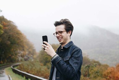 A man in eyeglasses standing on a road smiling looking at his smartphone.