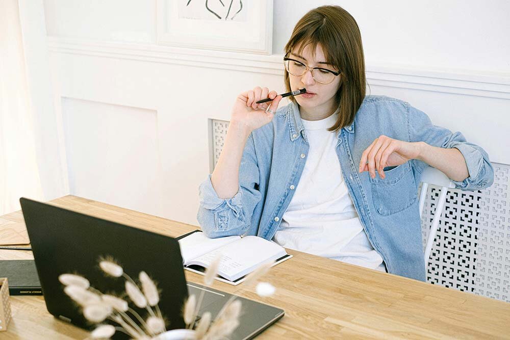 Young woman in denim shirt using computer and looking undecided.