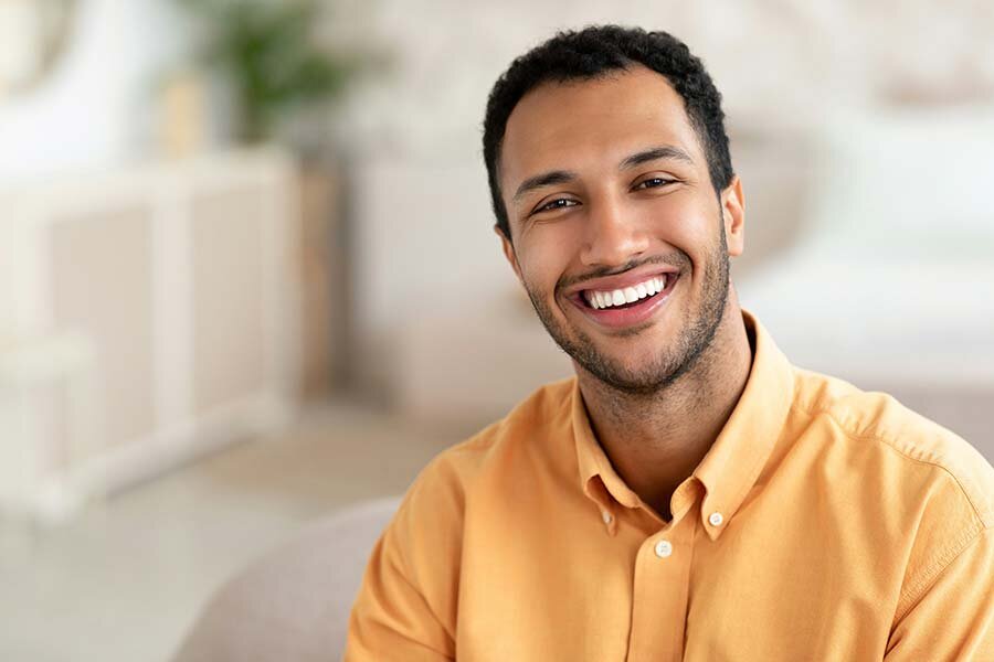 A smiling man sits comfortably on a grey couch in a bright living room.
