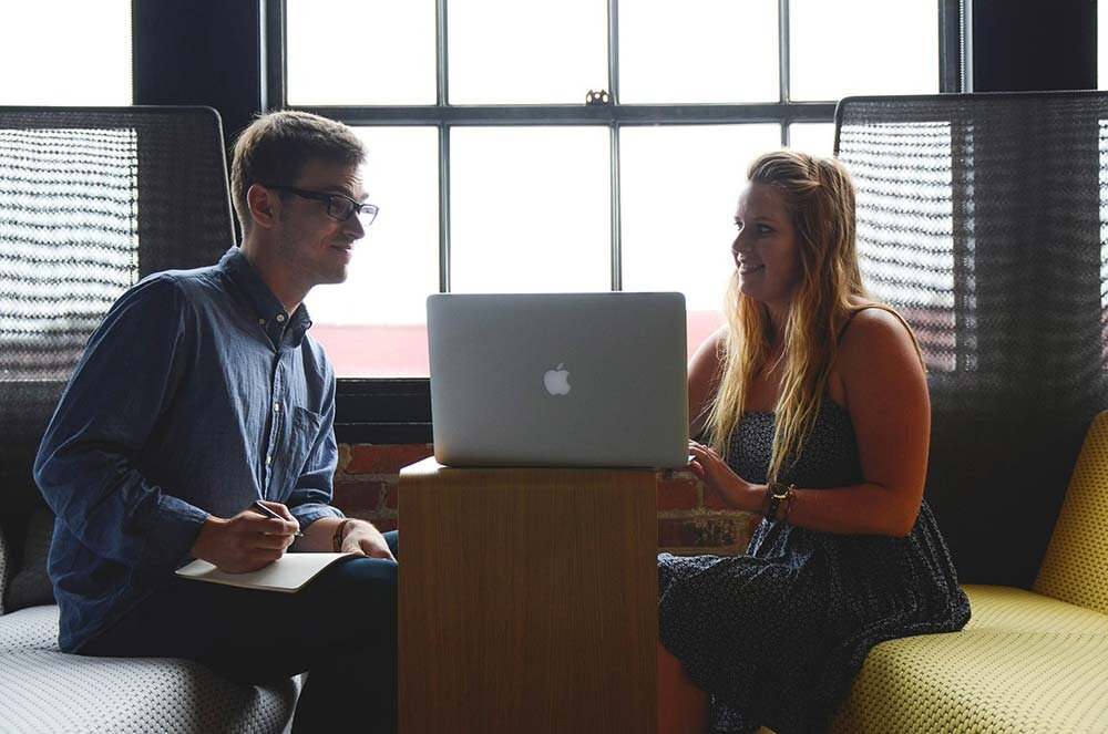Young man holding a pen with paper on his lap sitting across the table from a young lady talking to him about ideas from a laptop