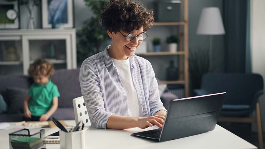 Woman sitting at a desk using a laptop with a child seated on a sofa in the background