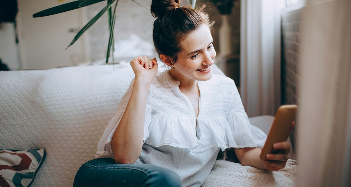 A woman seated on a white couch next to a window using a cell phone