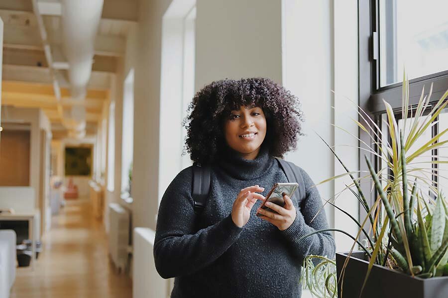 Woman in a dark long sleeve shirt standing next to a plant by the window using a smartphone to apply for a fixed-interest loan.