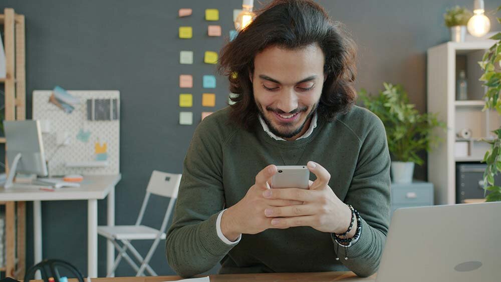 A young man seated at a desk, smiling while using his smartphone to apply for a loan.
