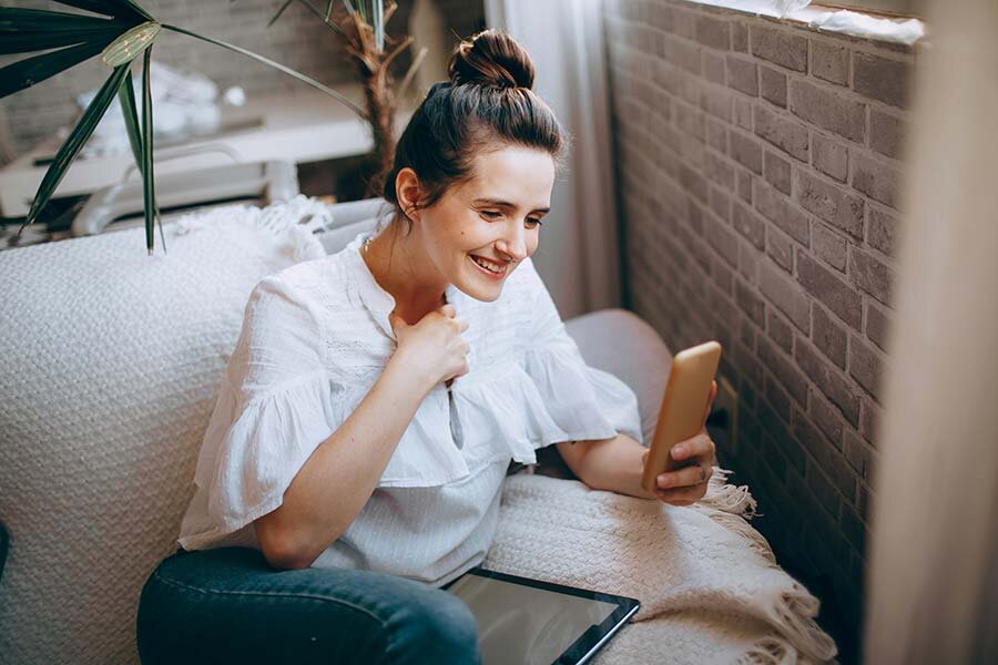 A woman sitting on a white sofa next to a brick wall using a smartphone.
