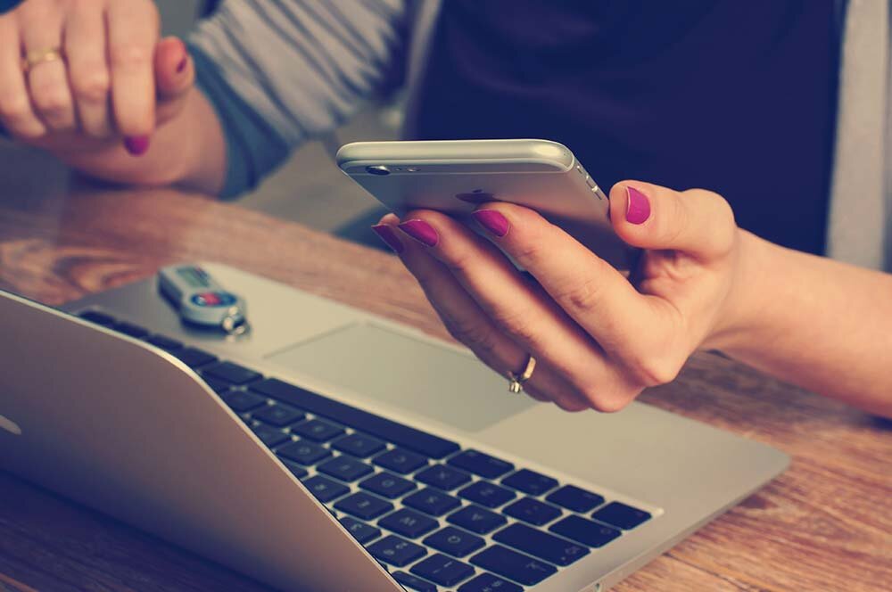 A woman sitting at a desk using a smartphone with a laptop computer in front of her