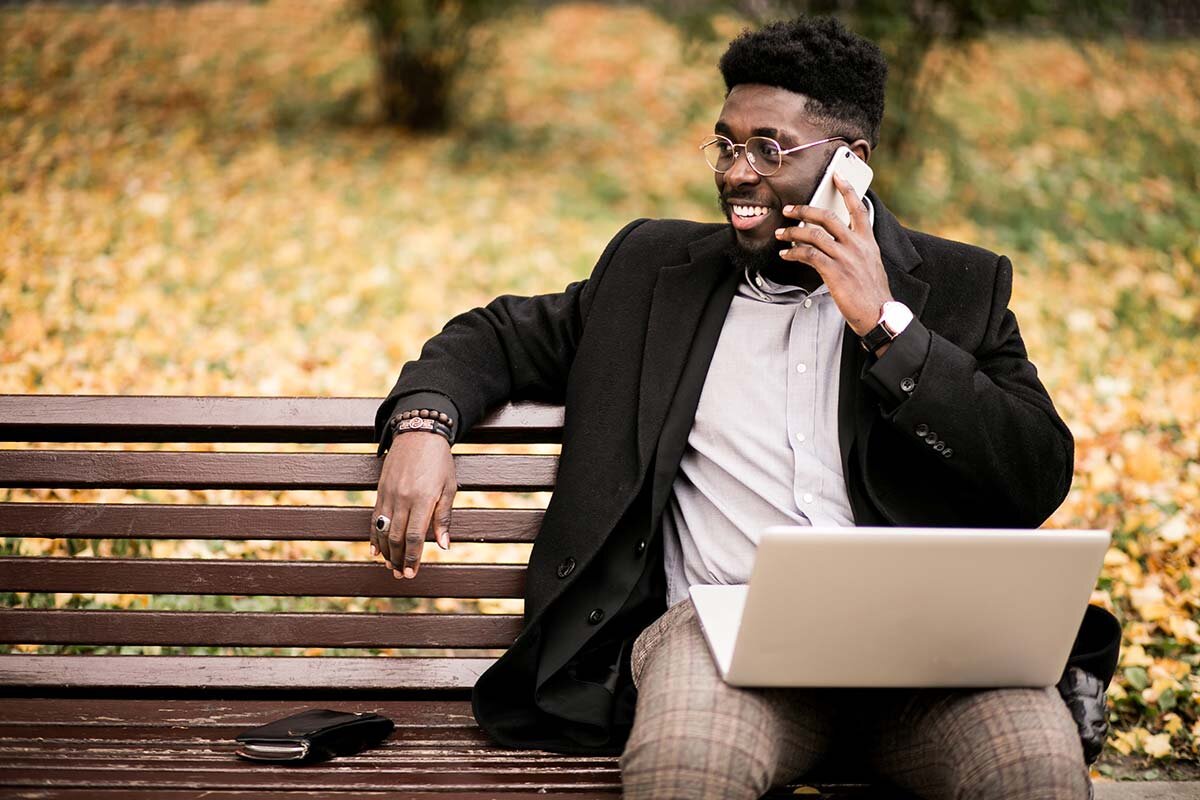A black man sitting on a park bench with a laptop on his lap and talking on his phone