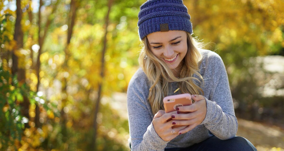 A woman sitting in the woods wearing a knit had holding a smartphone