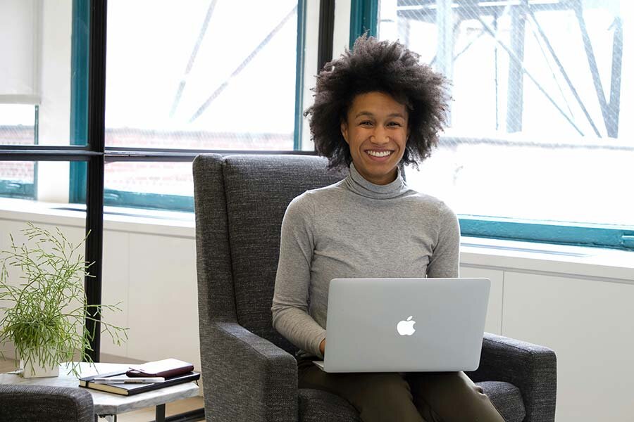 A smiling woman sitting on a gray chair using a MacBook to withdraw funds from a credit line.