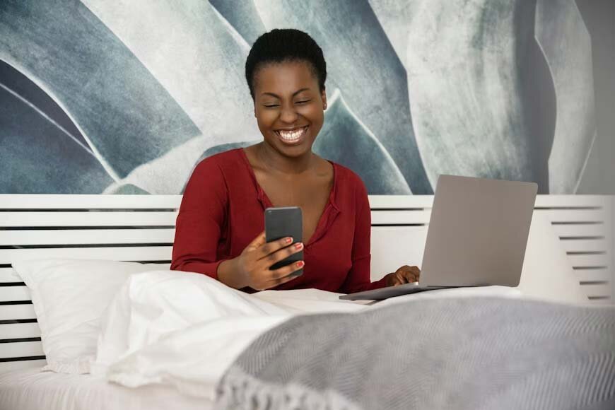A woman sitting in a bed with an open laptop and looking at her phone 