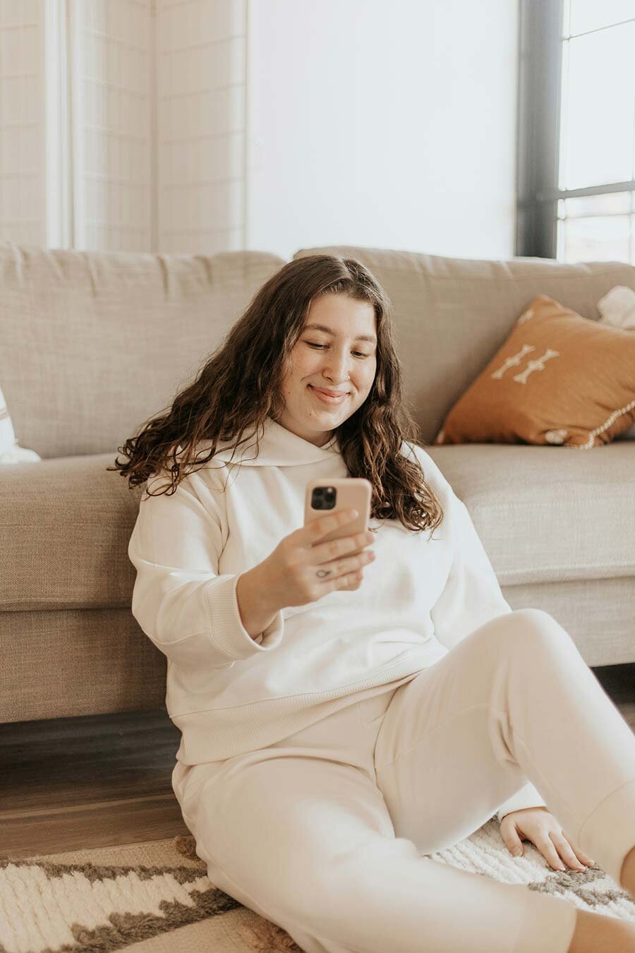 A woman seated on a carpeted floor with back leaning against a couch, using a cell phone